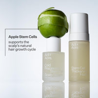 Two Act+ Acre. hair care bottles on a light gray background: a frosted bottle labeled Scalp Serum with a sliced green apple resting on top, and a taller bottle labeled Stem Cell Thickening Foam with pale yellow liquid inside.