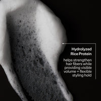 Close-up of white shampoo lather applied to dark hair against a black background, with text reading 'Hydrolyzed Rice Protein' and a descriptive callout.