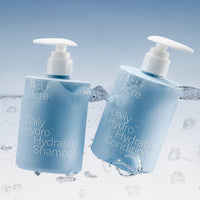 Two bottles of Act+Acre™ Daily Hydro Hydrating Shampoo and Conditioner with water droplets on a light blue background.