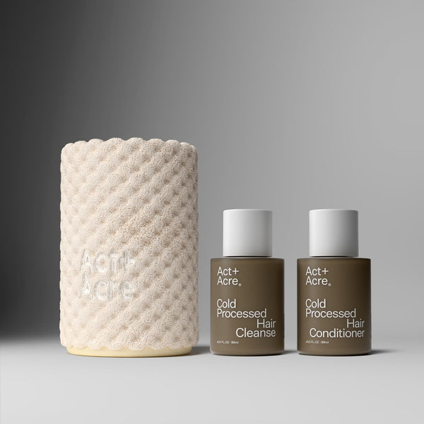 Two bottles of Act+Acre Cold Processed Hair Cleanse and Conditioner next to a textured beige container on a gray background.